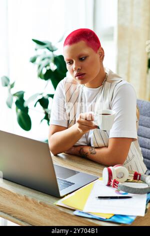 Eine junge Frau mit lebhaften Haaren trinkt Kaffee und konzentriert sich auf ihren Laptop in einem gemütlichen Arbeitsbereich. Stockfoto