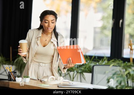 Die junge Frau konzentriert sich auf ihre Aufgaben, während sie in einem hellen Büro einen Kaffee hält und Dokumente hält. Stockfoto