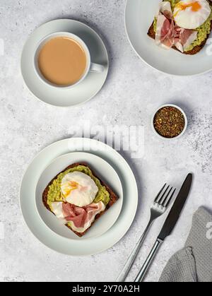 Lebensmittelfotografie von nahrhaftem Frühstück, Sauerteig-Roggenbrot mit Avocadopüree, Schinken, Speck, gekochtem pochiertem Ei, Tee mit Milch Stockfoto