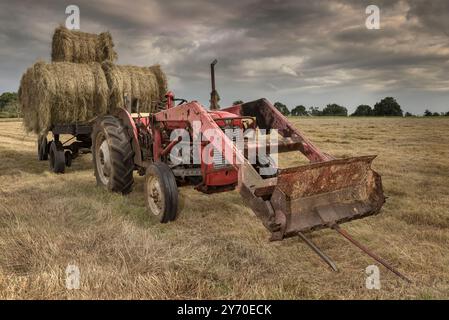 Bei der Ernte von Heuballen wird in Wiltshire, England, ein klassischer Traktor verwendet, wenn sich Sturmwolken nähern. Stockfoto