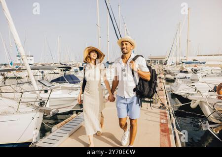 Ein lächelndes Paar spaziert Hand in Hand am Dock des Yachthafens entlang, umgeben von Booten an einem sonnigen Tag. Stockfoto