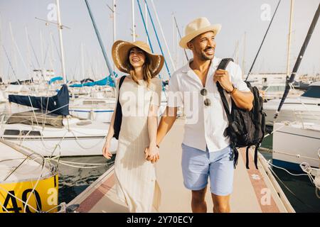 Ein lächelndes Paar spaziert Hand in Hand am Dock des Yachthafens entlang, umgeben von Booten an einem sonnigen Tag. Stockfoto