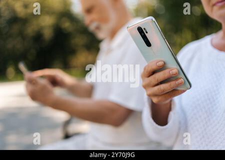 Close-up Porträt eines schönen älteren Paares mit Smartphones, die an sonnigen Sommertagen auf der Parkbank sitzen und in Verbindung bleiben Stockfoto