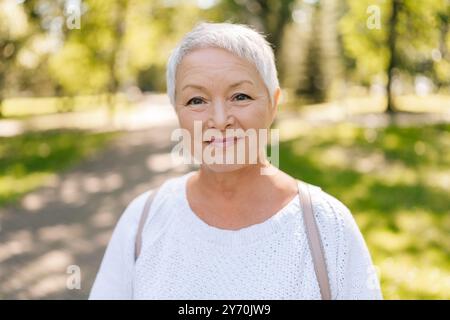 Porträt einer schönen grauhaarigen Seniorin, die in die Kamera blickt, die am sonnigen Tag im grünen Sommerpark posiert und Selbstvertrauen ausstrahlt Stockfoto