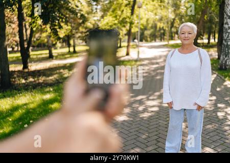 Selektive Fokusaufnahme einer grauhaarigen Seniorin, die sich glücklich im sonnigen Park posiert, während Ehemann Fotograf Momente mit Smartphone festnimmt. Stockfoto