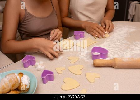 Cookies Baking Kitchen: Zwei Frauen machen in einer Küche herzförmige Kekse, indem sie Keksschneider und Teig auf einer bemehlten Oberfläche verwenden. Stockfoto