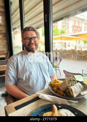 Millennial Mann mit Brille und Bart sitzt an einem Restauranttisch und genießt eine Mahlzeit. Entspannte Atmosphäre und Stadtleben, sichtbar am Fenster. Cafe Stockfoto