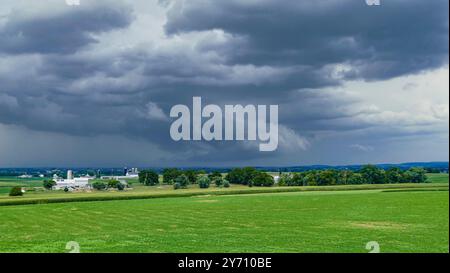 Dunkle Wolken erheben sich auf unheilvolle Weise über einem riesigen grünen Feld und deuten auf einen herannahenden Sturm hin. In der Ferne kann man einen Bauernhof sehen, der von der üppigen Landschaft eingerahmt wird, während der Tag in die Abenddämmerung geht. Stockfoto