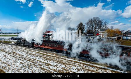 Eine alte Dampfeisenbahn bewegt sich durch eine Landschaft aus schneebedeckten Feldern unter einem klaren blauen Himmel und weht weißen Dampf, während sie durch die Landschaft fährt. Stockfoto