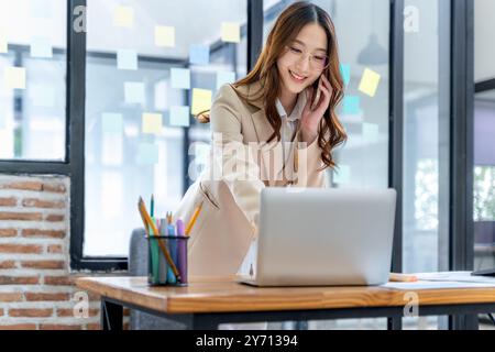 Glückliche junge Geschäftsfrau, die allein im Büro an ihrem Projekt arbeitet und mit ihrem Kunden am Telefon spricht Stockfoto
