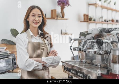 Glückliche junge weibliche Coffee-Shop-Besitzerin, die hinter ihrer Theke neben ihrer Kaffeemaschine steht und lächelt Stockfoto