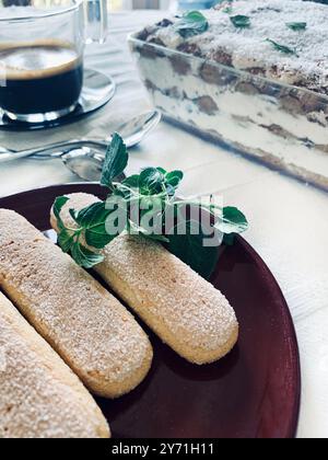 Ladyfinger-Kekse für die Zubereitung von Tiramisu auf einem Teller, auf verschwommenem Hintergrund ein Tablett mit Tiramisu und Kaffee. Italienisches Tiramisu-Konzept. Stockfoto