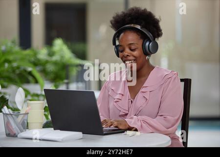 Mittelgroße Aufnahme einer lächelnden afroamerikanischen Geschäftsfrau, die am Tisch sitzt und mit einem Laptop an einem Online-Meeting teilnimmt, während sie ferngesteuert vom Café aus arbeitet Stockfoto