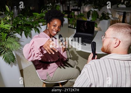 Fröhliche weibliche Podcast-Moderatorin der afroamerikanischen Ethnicity winkte spielerisch die Hand zur Kamera und nahm eine neue Episode mit männlichem Gast am Tisch im Café mit grünen Pflanzen und Kamerablitz auf Stockfoto