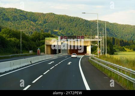 Ein einsames Fahrzeug nähert sich dem Tunneleingang, umgeben von üppigen grünen Hügeln unter einem hellblauen Himmel, und lädt zu einer friedlichen Reise auf einem sonnigen af ein Stockfoto