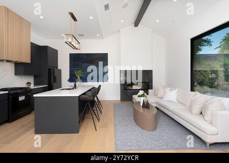 Moderne offene Küche und Wohnzimmer mit eleganten schwarzen Schränken, weißen Arbeitsflächen und einem gemütlichen Sitzbereich mit großem Fenster. Stockfoto