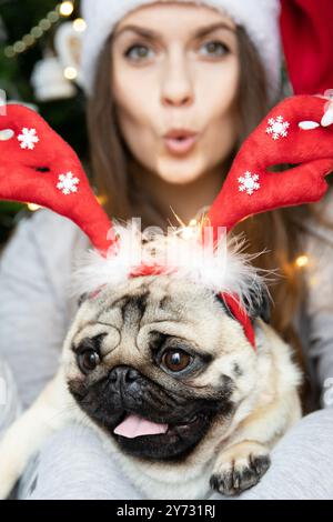 Lustiger süßer Mops Hund in roten Weihnachts-Rentier Hörner, der in den Armen eines schönen Mädchens mit Santa Hut sitzt Stockfoto