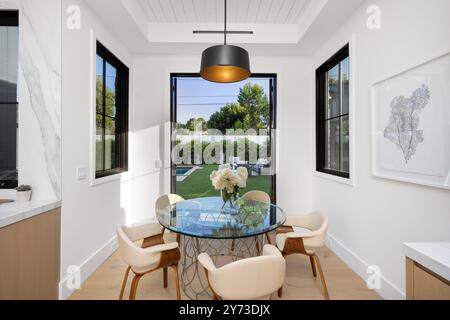 Moderner Essbereich mit Glastisch, weißen Stühlen und großen Fenstern mit Blick auf den begrünten Hinterhof. Stockfoto