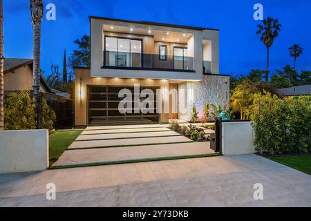 Ein modernes zweistöckiges Haus mit einer gut beleuchteten Außenanlage und einer Auffahrt, umgeben von Palmen und Grün in der Abenddämmerung. Stockfoto
