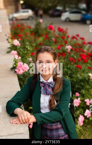 Mädchen in Schuluniform, auf einem Felsvorsprung gestützt, umgeben von blühenden Blumen, selbstbewusst lächelnd. Straßenporträt eines Teenager-Mädchens Stockfoto