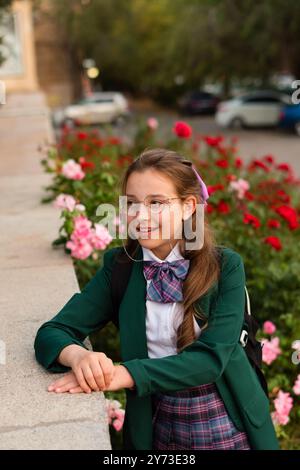 Mädchen in Schuluniform, auf einem Felsvorsprung mit einem sanften Lächeln, umgeben von blühenden Blumen. Straßenporträt eines Schulmädchens Stockfoto
