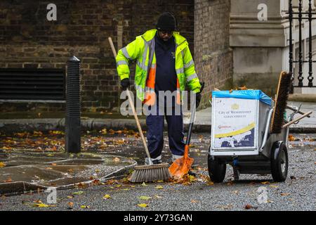 London, Großbritannien. September 2024. Ein Mitarbeiter des Westminster council fegt fleißig die Straße und den Straßenbelag in der Downing Street und sammelt Haufen von bunten, gefallenen Blättern. An einem dezidiert herbstlichen Tag im Zentrum Londons wurden heute starke Schauerwellen, Blauwind und kältere Temperaturen beobachtet. Quelle: Imageplotter/Alamy Live News Stockfoto