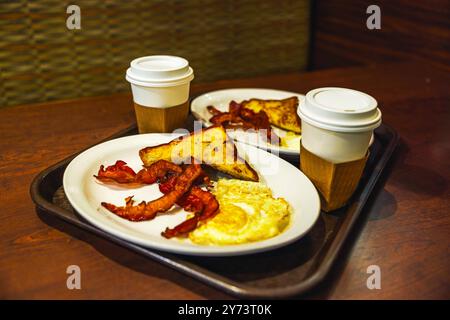 Teller mit Speck, Spiegeleiern, Toast und Kaffeetassen auf einem Holztisch in einem Café am Flughafen Miami. Stockfoto