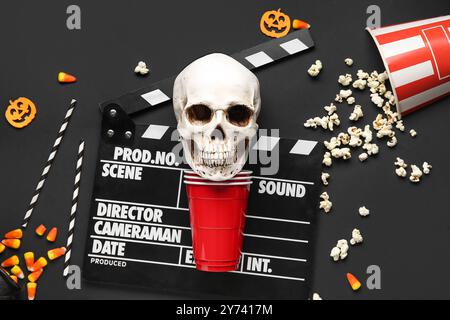 Komposition mit Klapperbrett, Plastikbechern, Leckereien und menschlichem Schädel für Halloween auf dunklem Hintergrund Stockfoto