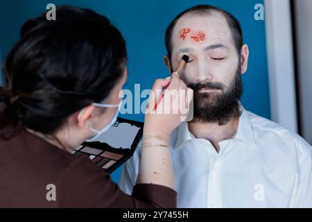 Freundin, die professionelles Augen-Make-up auf den Mann aufträgt, damit er tot und krank für halloween aussieht und ihn als Zombie verkleidet. Frau, die die Grundlage für blasses Gesicht und Narben auf ihrem Freund aufträgt Stockfoto