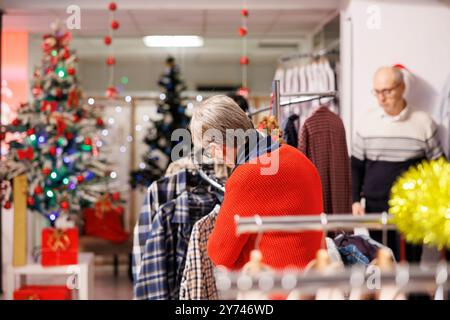 Ältere Frau, die während des saisonalen Verkaufs im Einzelhandel durch Kleidung stöbert und in der Boutique nach Weihnachtsessen mit Dekorationen sucht. Ein älterer Kunde im Einkaufszentrum kauft Geschenke. Stockfoto