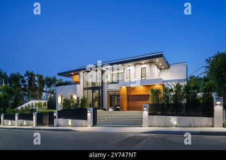 Modernes Luxushaus mit großen Fenstern, beleuchtet in der Abenddämmerung, umgeben von Grün. Stockfoto