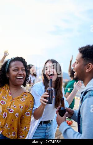 Drei verschiedene junge Freunde tanzen zusammen und halten Bierflaschen auf einer Sommerparty auf dem Dach Stockfoto