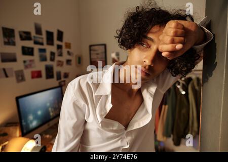 Ein kranker Kerl in weißem Hemd, der durch das Fenster schaut, während er an der Wand steht und an Kater leidet Stockfoto