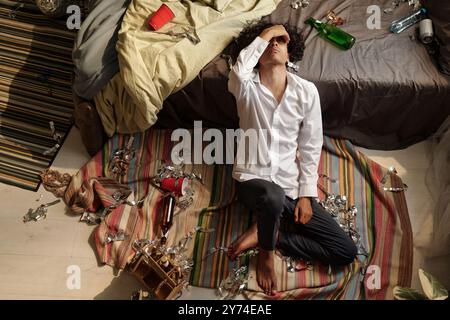 Hoher Winkel eines jungen Mannes, der an einem Kater leidet, der seinen Kopf berührt und ihn am Bettrand hält, während er auf dem Boden sitzt Stockfoto