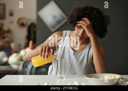 Junger Mann mit Kater-Symptomen, der am Tisch sitzt und Orangensaft in Glas gießt, während er nach Alkohol frühstückt Stockfoto