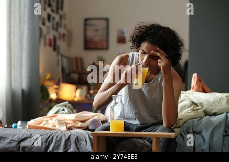 Ein Teenager mit Alkoholvergiftung sitzt auf dem Bett in einem unordentlichen Zimmer und trinkt nach der Heimfeier ein Glas Orangensaft Stockfoto