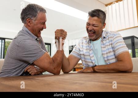 Armwrestling und Lachen, zwei ältere verschiedene Freunde genießen die gemeinsame Zeit drinnen, zu Hause Stockfoto