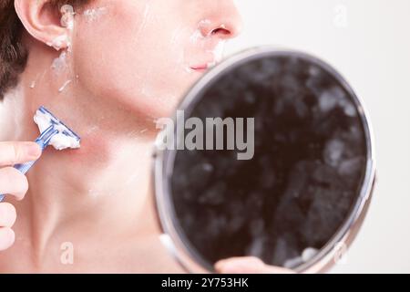 Junger Mann rasiert sich vorsichtig mit einem blauen Rasierer im Badezimmerspiegel und konzentriert sich auf die Pflege Stockfoto