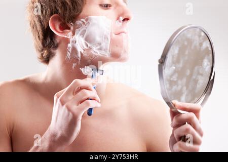 Im Badezimmer schaut ein junger Mann vorsichtig in den Spiegel, während er sein Gesicht mit einem blauen Rasierer und einer Rasiercreme rasiert, wobei er sich auf seine Hygiene und seine Pflege konzentriert Stockfoto