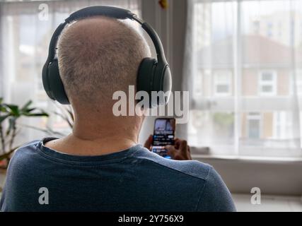 Ein älterer Mann, der sich zu Hause entspannt und Online-Musik mit schnurlosen Kopfhörern und einem Smartphone hört. Ein Konzept der Technologienutzung. Stockfoto