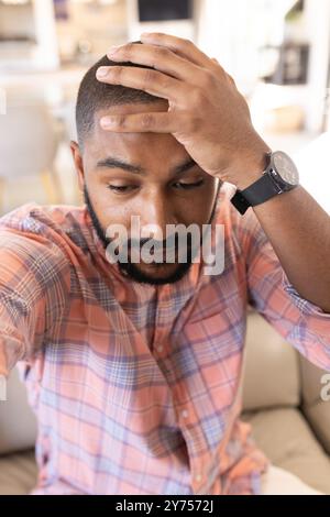 Auf der Couch sitzend, indischer Mann in kariertem Hemd, der den Kopf hält und gestresst aussieht, während eines Videogesprächs Stockfoto