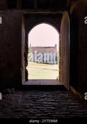 Ein Blick aus dem Inneren eines alten Steingebäudes mit Blick durch eine bogenförmige Tür auf ein traditionelles Landhaus mit grünem Rasen. Stockfoto