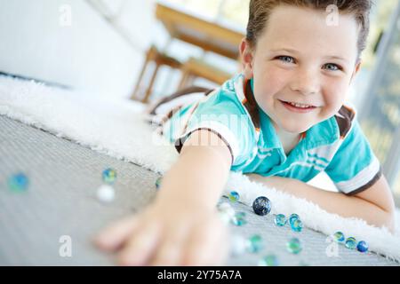 Fröhliche, Porträts- und Kinderspiele mit Marmor auf dem Boden, um lustige Spiele zu spielen oder zu Hause zu entspannen. Gesicht, Lächeln und süßer Junge mit Glaskugeln oder Spielzeug auf Teppich für Stockfoto