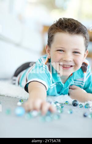 Lächeln, Portrait und Kinderspiel mit Marmor auf dem Boden für lustige Spiele, Entspannung oder Lachen zu Hause. Gesicht, glücklicher Junge und süßes Kind mit Glaskugeln oder Spielzeug Stockfoto