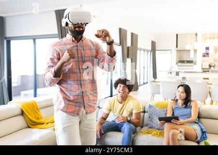Mit dem VR-Headset spielt ein Mann virtuelles Spiel, während verschiedene Freunde zuhause zusehen und lachen Stockfoto