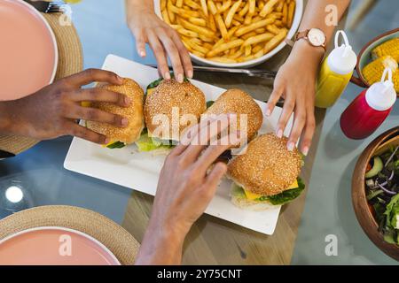 Verschiedene Freunde greifen nach Burgern und Pommes Frites und genießen gemeinsam am Tisch, zu Hause Stockfoto