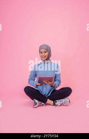 Eine junge Frau in einem blauen Pullover sitzt entspannt mit einem Tablet vor rosa Hintergrund Stockfoto