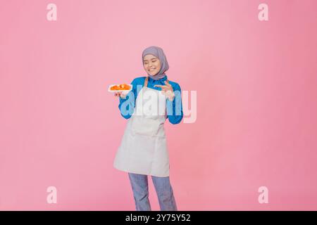 Eine fröhliche junge Frau in einer stilvollen Schürze hält eine Platte mit bunten Früchten vor einem rosafarbenen Hintergrund Stockfoto