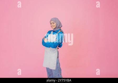 Eine fröhliche Frau in stilvoller Schürze und Hijab posiert anmutig vor einem rosa Hintergrund Stockfoto