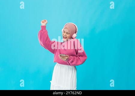 Eine junge Frau tanzt fröhlich in stilvollen Kopfhörern vor einem leuchtend blauen Hintergrund und verkörpert Spaß Stockfoto